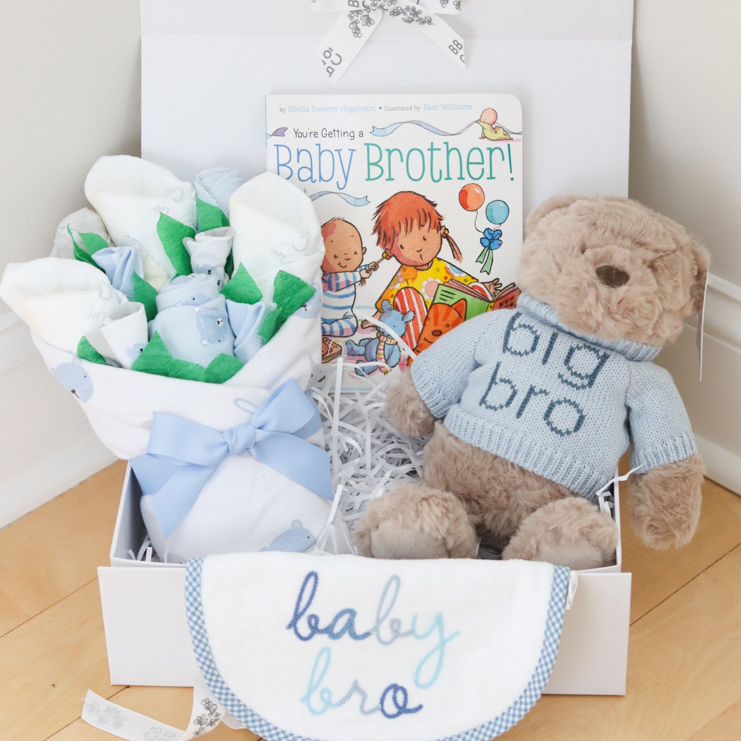 Baby gift set for big brother and new baby brother with teddy bear, baby clothes, and book on a wooden floor.