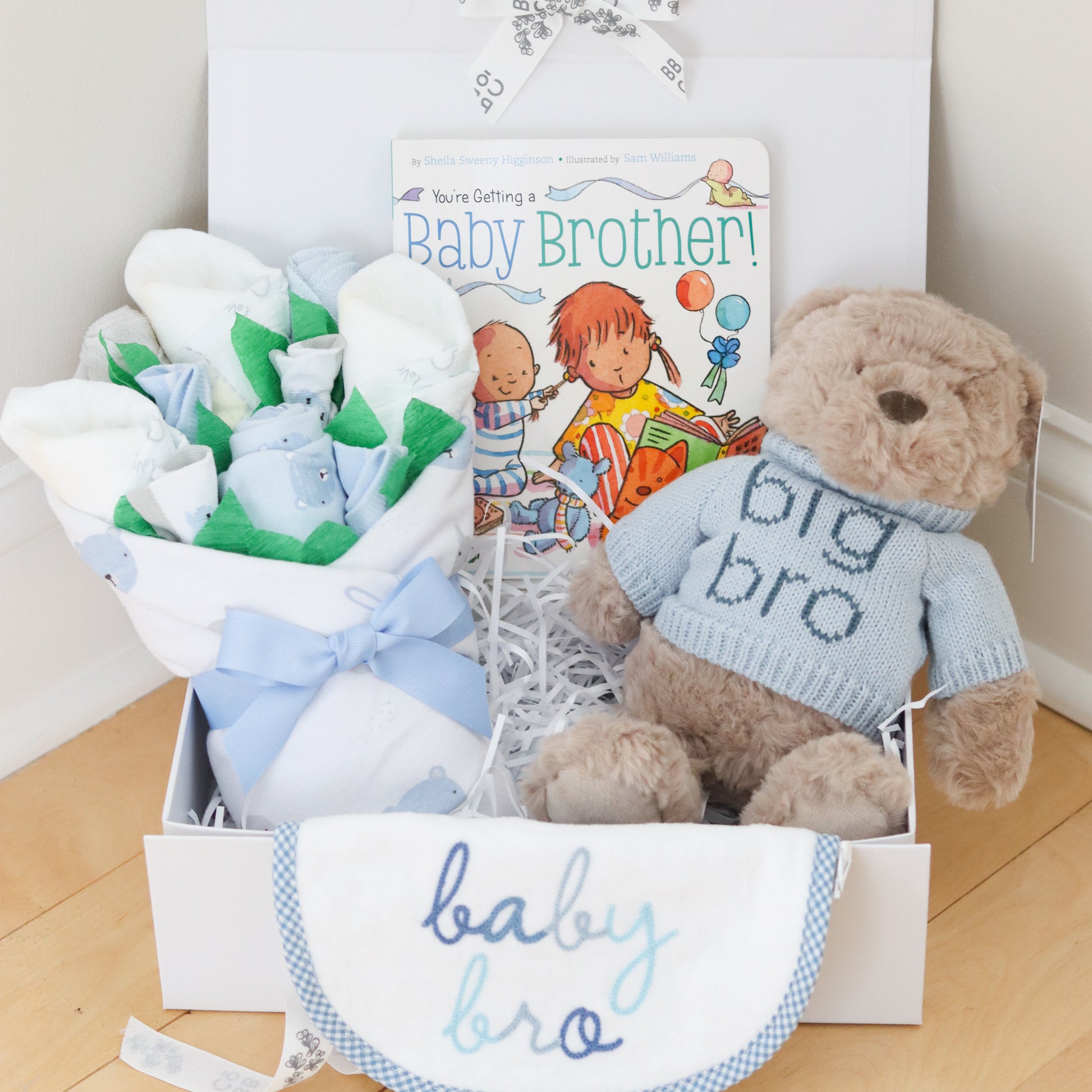Baby gift set for big brother and new baby brother with teddy bear, baby clothes, and book on a wooden floor.