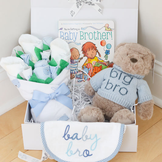 Baby gift set for big brother and new baby brother with teddy bear, baby clothes, and book on a wooden floor.