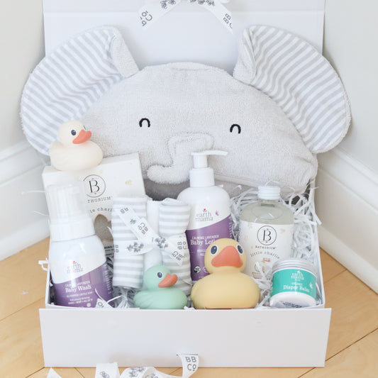 Baby care products including bath wash, lotion, washcloths, and rubber ducks in a box with an elephant design on a light wooden floor.