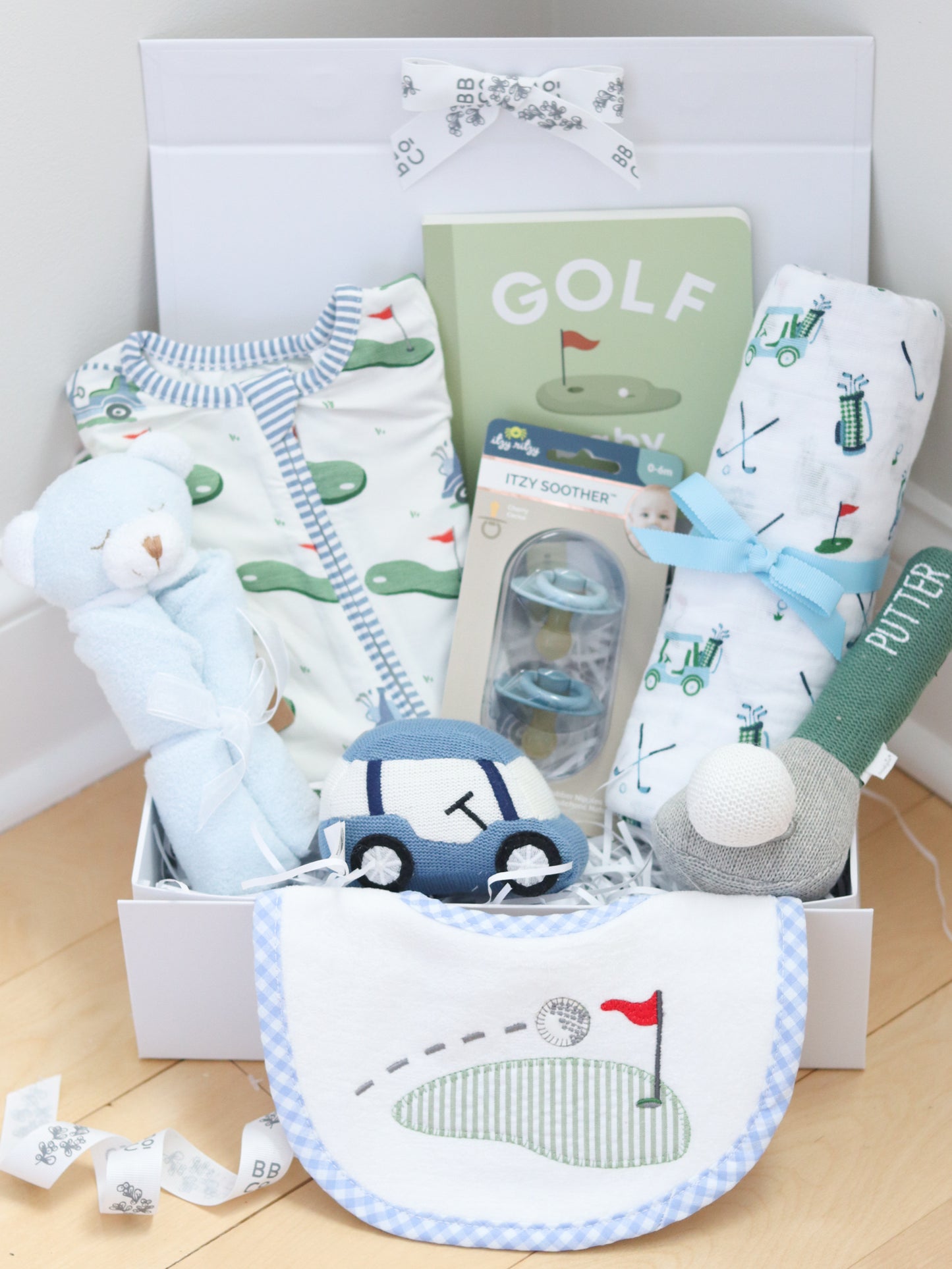 Baby gift basket with baby clothes, bib, and toys on a wooden floor.