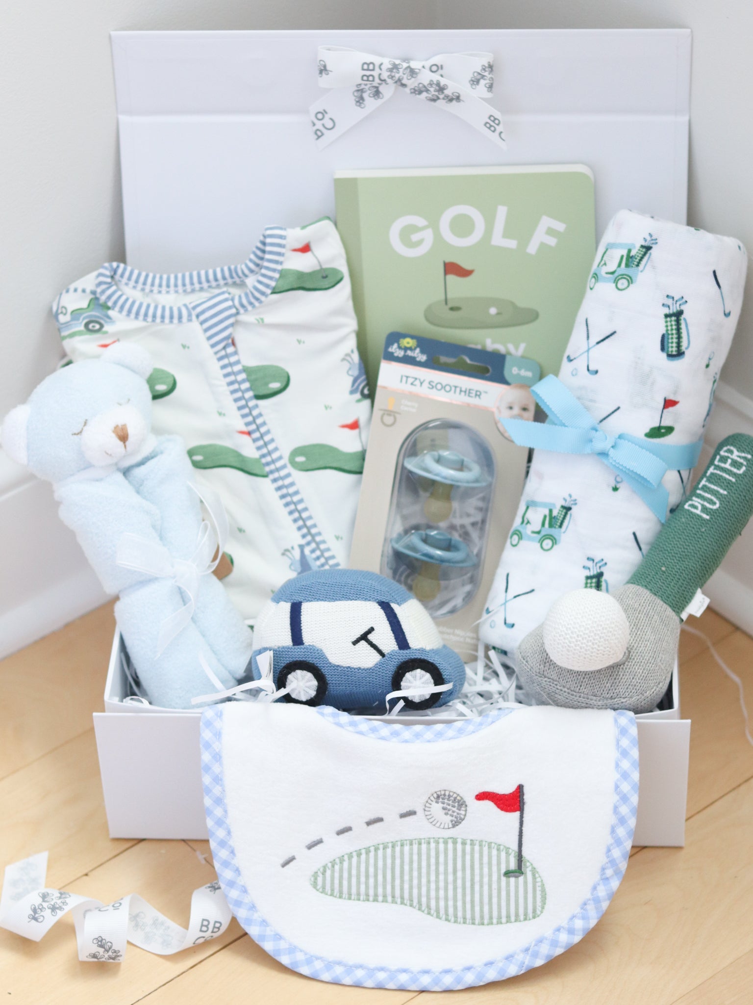 Baby gift basket with baby clothes, bib, and toys on a wooden floor.