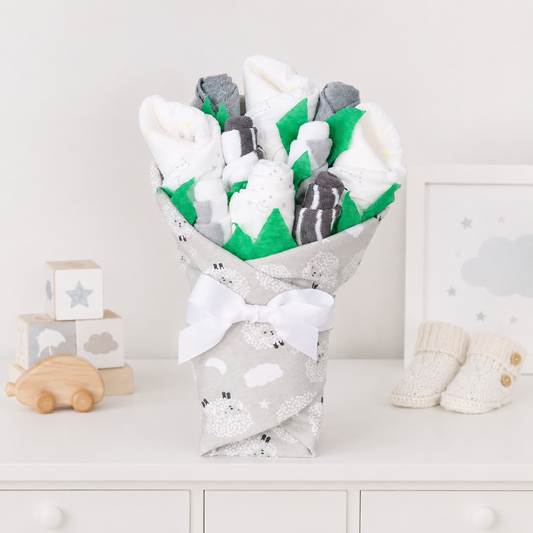 A baby gift bouquet styled like a fabric flower arrangement, containing white and gray knit socks and a white blanket, wrapped in a gray sheep-print fabric with a white bow, displayed on a white dresser with baby props.