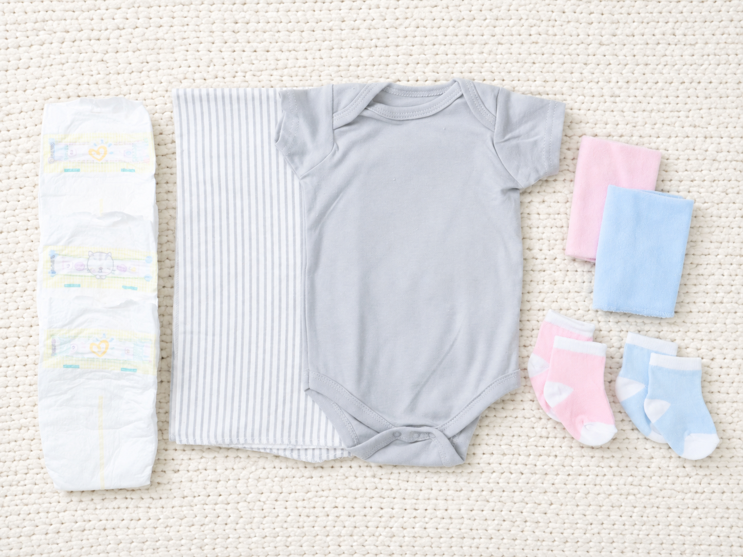 Baby clothing and diaper set laid flat on a textured cream woven rug, including a light gray short-sleeve bodysuit, a striped diaper cover, a white diaper, folded pink and blue socks, and folded pink and blue washcloths.