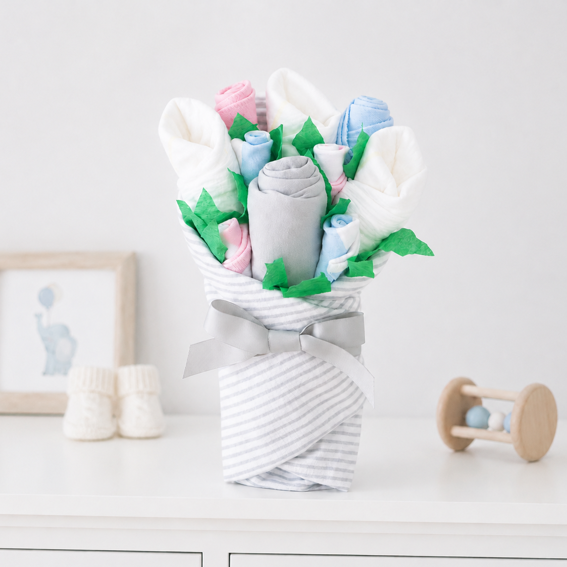 A baby gift bouquet made from folded white, pink, blue, and gray blankets or swaddle fabrics, arranged with green tissue paper leaves and wrapped in a white-and-gray striped fabric with a gray ribbon bow, displayed on a white dresser.