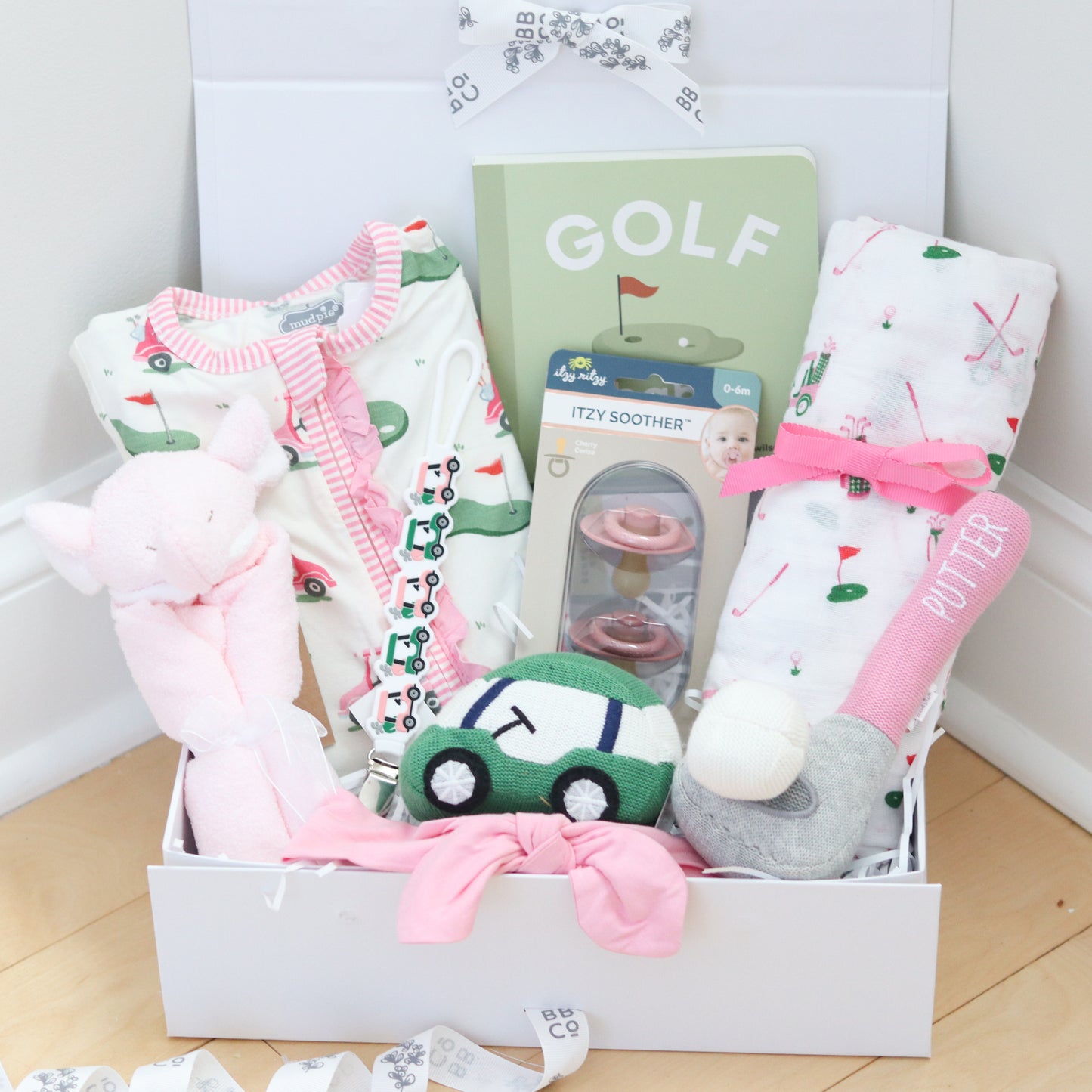 Golf baby girl gift box with baby clothes, toys, and a book on a wooden floor.