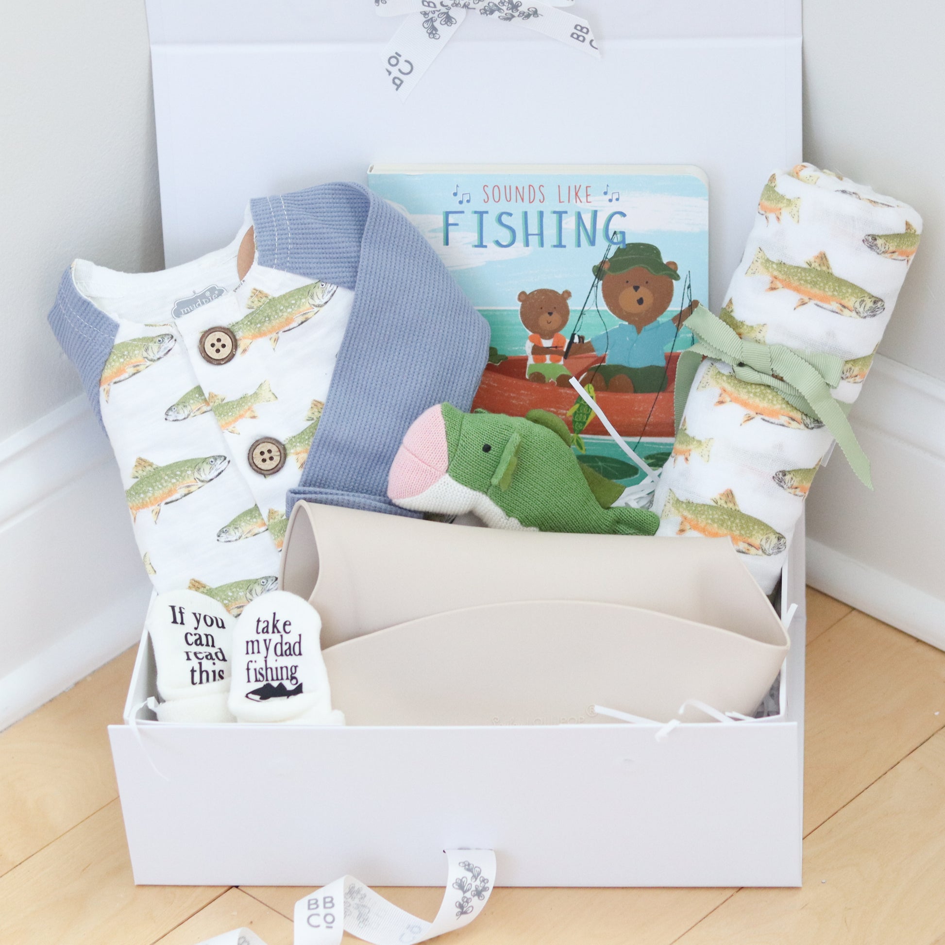 Fishing theme baby gift set with baby items including a blanket, book, and socks on a wooden floor.