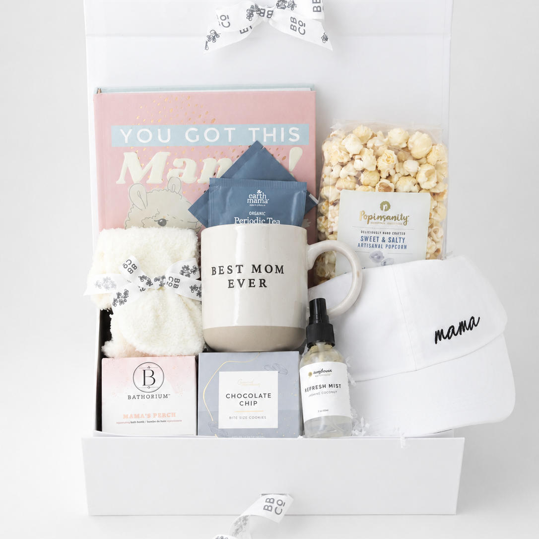 A curated gift box set fora new Mom, featuring a white ceramic mug with 'BEST MOM EVER' printed on it, a pink 'YOU GOT THIS Mom' book, a blue bag of tea, a box of bath bombs, a box of chocolate chip cookies, a bottle of refresh mist, a white cap with 'mama' written on it, a bag of popcorn all arranged in a white gift box with a white ribbon.