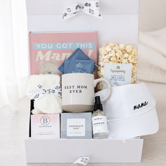 A curated gift box set for moms, containing a white ceramic mug that reads 'BEST MOM EVER', a bagof popcorn, a small bottle of refresh mist, tea packets, a white cap embroidered with 'mama', and other gift items, all arranged inside a white box with a white ribbon bow.